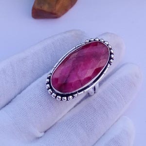 May include: An oval-shaped ring featuring a large, faceted, deep pink gemstone set in a silver band. The setting is decorated with a beaded border around the edge of the stone, and the metal band displays a textured, oxidized finish along the sides.