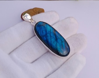 May include: A silver pendant with a large, oval labradorite stone. The stone has a vibrant blue color with streaks of darker blue and black. The pendant is held by a silver bail.