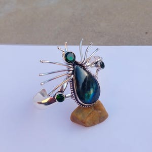 以下が含まれることがあります： 大きな洋梨型の虹色の宝石をあしらったシルバーリング。リングは、複数の緑色の宝石と、中央の石から伸びる繊細な金属細工が特徴的なユニークなデザインです。リングは、小さな茶色の岩の上に展示されています。
