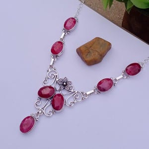 May include: A silver necklace featuring seven oval-shaped, faceted deep pink ruby-colored stones set in metal bezels. The central design includes a small silver flower accent surrounded by intricate silver scrollwork connecting the stones.