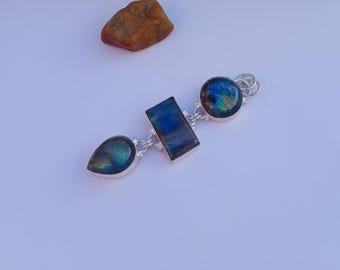 May include: A silver pendant featuring three labradorite stones in a teardrop, rectangular, and circular shape. The stones display blue and green iridescence. A small, brown rock is in the background.