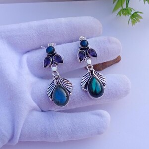 Puede incluir: Un par de pendientes plateados con diseño de hojas. Cada pendiente presenta una piedra azul-verdosa iridiscente en forma de lágrima y piedras moradas y azules más pequeñas. Los pendientes se exhiben sobre una superficie blanca.