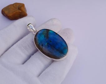 May include: Oval-shaped pendant with a vibrant blue and green gemstone set in a silver bezel. The gemstone has a shimmering, iridescent quality. The pendant is held by a gloved hand, with a small, brown rock in the background.