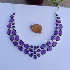 Peut inclure: Un collier en argent orné de multiples pierres précieuses améthystes violettes de forme ovale et en goutte d'eau, disposées en un motif en croissant à double rangée. Le bijou comprend une chaîne en métal et le texte Khushi artisan sur la droite.
