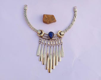 May include: A gold-coloured necklace featuring a blue gemstone pendant and hanging teardrop charms. The necklace has a textured chain and is displayed on a light purple surface. A small, brown stone sits above the necklace.