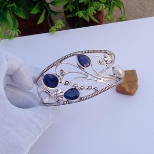 May include: A silver-toned cuff bracelet with an openwork design. The bracelet features several dark blue gemstones and a single white gemstone. The bracelet has an ornate, floral-inspired pattern.