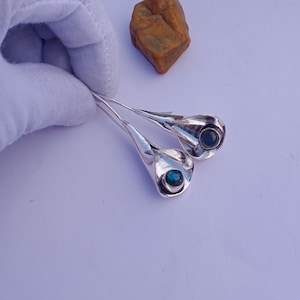 May include: Silver-toned calla lily earrings with a blue gemstone center. The earrings have a sleek, modern design, with a curved, elongated shape. The gemstones are a deep blue color, adding a pop of color to the earrings. The earrings are displayed on a light purple surface.