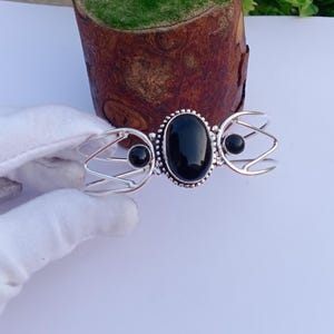 Könnte beinhalten: Silberfarbenes Armband mit einem großen, ovalen schwarzen Edelstein in der Mitte, flankiert von kleineren schwarzen Steinen. Das Armband hat ein offenes Design mit filigranen Drahtarbeiten auf beiden Seiten des zentralen Steins. Der Hintergrund ist neutral.