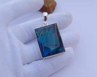 May include: A rectangular labradorite pendant with a silver-colored frame. The stone displays iridescent blue and green hues. The pendant is held by a gloved hand.