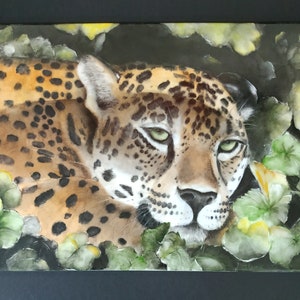 May include: A close-up of a jaguar's face, with green eyes, lying in a bed of green leaves. The jaguar's fur is brown and black with spots.