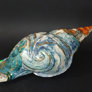 May include: A large seashell sculpture painted with swirling blue, white, and gold patterns. The shell is decorated with a detailed design of a wave and has a small orange and white section at the tip.