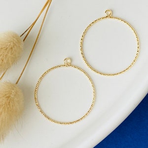 Puede incluir: Dos colgantes circulares de metal dorado texturizado con pequeños bucles en la parte superior para colgar. Los colgantes están sobre una superficie blanca con elementos decorativos beige secos y un fondo azul.