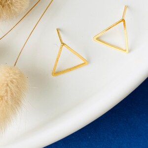 May include: Two gold-colored, open-frame triangle earrings. The earrings are displayed on a white surface with dried, fluffy beige plants. The background is a deep blue.