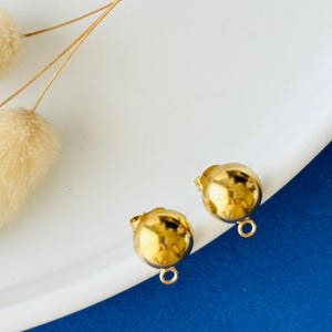 Op de afbeelding: Twee glanzende, goudkleurige kogelvormige oorbellen met kleine lusjes aan de onderkant. De oorbellen liggen op een wit oppervlak met een blauwe accent. Gedroogde, pluizige beige bloemen zijn op de achtergrond.