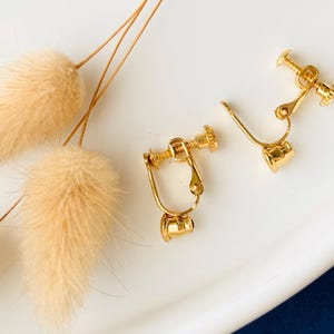 Könnte beinhalten: Nahaufnahme von zwei goldfarbenen Clip-Ohrringen mit schraubenähnlichem Design und einer einzelnen Ohrringkomponente. Die Ohrringe liegen auf einer weißen Oberfläche, mit getrockneten, flauschigen, beigefarbenen Dekorationselementen.