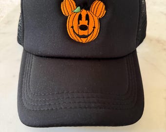 May include: A black trucker hat featuring an embroidered orange pumpkin shaped like a mouse head on the front panel. The pumpkin design includes a small green leaf detail, black stitched facial features, and vertical lines to mimic the texture of a gourd.