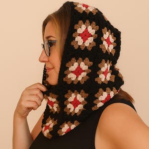May include: A black crochet hood with a floral pattern in red, brown, and cream. The hood is made of square crochet pieces and is worn around the neck and head. The hood is a handmade item.