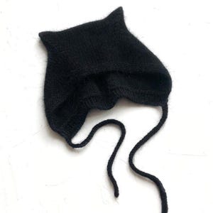 May include: A black knitted baby bonnet with pointed ears and long ties. The bonnet is made of a soft, textured yarn and is designed to cover the head and neck. The ties are used to secure the bonnet under the chin.