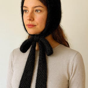 May include: A black knitted bonnet with long ties, tied in a bow under the chin. The bonnet is made of a soft, textured yarn. The person in the image is wearing a light beige sweater.