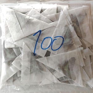 May include: A pile of 100 white paper tea bags with a dark green herbal tea inside. The tea bags are folded into triangles and are stacked on top of each other.