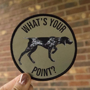 May include: A round, green and black patch with a black and white dog silhouette and the text "WHAT'S YOUR POINT?"