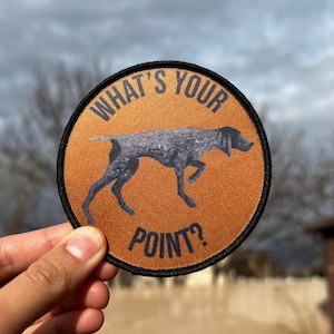 May include: A round, brown patch with black stitching features a black and white illustration of a dog pointing. The text "What's Your Point?" is printed in black on the patch.