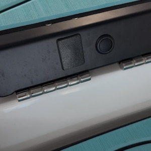 May include: Close-up of a teal and black boat deck with a black panel featuring a small black button. The panel is attached to a white surface with a row of silver metal hinges.