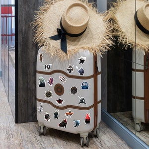May include: A gray suitcase with a variety of stickers featuring characters and symbols from the popular fantasy series The Witcher. The stickers are arranged in a random pattern on the suitcase, which is standing upright on a wooden floor.