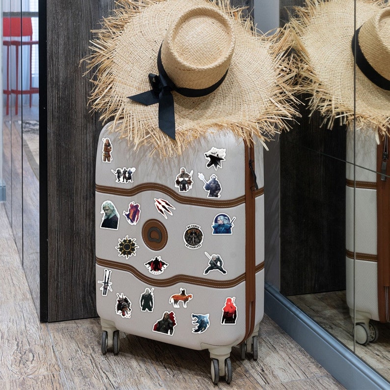 May include: A gray suitcase with a variety of stickers featuring characters and symbols from the popular fantasy series The Witcher. The stickers are arranged in a random pattern on the suitcase, which is standing upright on a wooden floor.