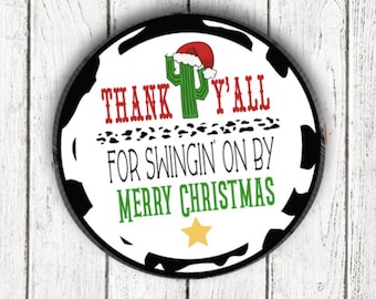 Könnte beinhalten: Runder Weihnachtsschmuck mit einem schwarz-weißen Kuhfellmuster-Rand. Der Schmuck zeigt einen grünen Kaktus mit Weihnachtsmütze und den Text "THANK Y'ALL FOR SWINGIN' ON BY MERRY CHRISTMAS" in Rot und Grün.
