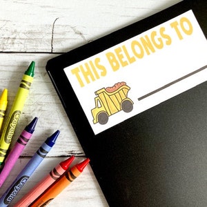 May include: A black notebook featuring a white rectangular label with the yellow text THIS BELONGS TO and a graphic of a yellow dump truck, accompanied by five colorful Crayola crayons in yellow, purple, blue, pink, and orange hues.