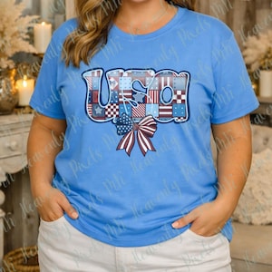 May include: A light blue t-shirt featuring the word "USA" in a patchwork design with red, white, and blue patterns. A bow with red and white stripes is below the word. The shirt is short-sleeved.