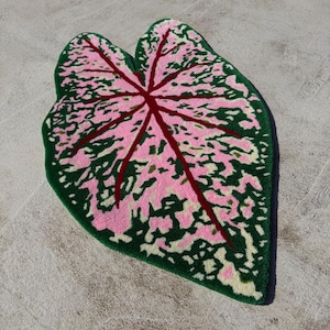 May include: A leaf-shaped rug with a vibrant design. The rug features a pink and green pattern, with a dark red vein structure. The edges are a deep green, and the overall shape mimics a large leaf. The rug is designed for home decor.
