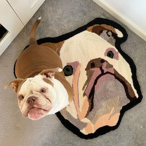 May include: A brown and white bulldog puppy looking up at the camera. The puppy is standing on a rug that is shaped like a bulldog's head. The rug is made of a soft, plush material and has a black outline.