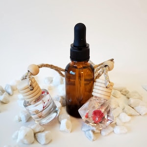 May include: An amber glass dropper bottle and two small, clear glass car air fresheners. The air fresheners are filled with dried flowers and have wooden tops. The bottles are displayed on white rocks.