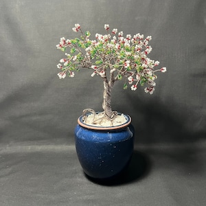 May include: A beaded wire tree with white and red flowers in a blue ceramic pot. The tree is made of wire and beads and is a decorative piece.