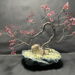 K&ouml;nnte beinhalten: Eine Drahtskulptur eines Bonsai-Baums mit rosa Bl&uuml;ten. Der Baum steht auf einem Steinsockel mit kleinen wei&szlig;en Steinen.