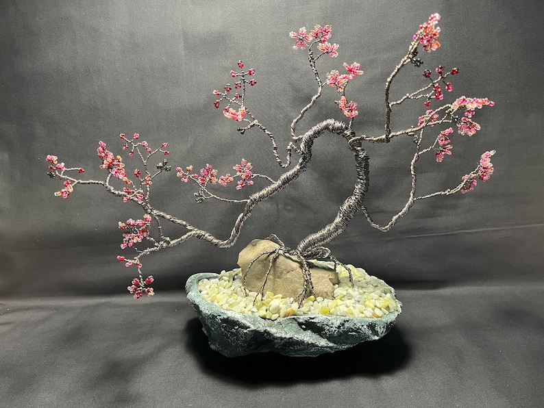 K&ouml;nnte beinhalten: Eine Drahtskulptur eines Bonsai-Baums mit rosa Bl&uuml;ten. Der Baum steht auf einem Steinsockel mit kleinen wei&szlig;en Steinen.