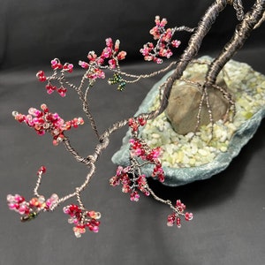 K&ouml;nnte beinhalten: Ein Bonsai-Baum aus Draht mit rosa und wei&szlig;en Perlen, die Bl&uuml;ten darstellen, und gr&uuml;nen Perlen, die Bl&auml;tter darstellen. Der Baum ist in einem grauen Steinsockel mit wei&szlig;en Kieselsteinen verwurzelt.