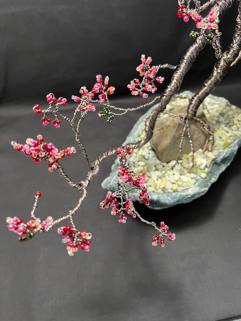 K&ouml;nnte beinhalten: Ein Bonsai-Baum aus Draht mit rosa und wei&szlig;en Perlen, die Bl&uuml;ten darstellen, und gr&uuml;nen Perlen, die Bl&auml;tter darstellen. Der Baum ist in einem grauen Steinsockel mit wei&szlig;en Kieselsteinen verwurzelt.