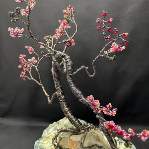 K&ouml;nnte beinhalten: Eine Drahtskulptur eines Bonsai-Baums mit rosa und gr&uuml;nen Perlen, die Bl&uuml;ten darstellen. Der Baum ist in einem grauen Steinsockel mit wei&szlig;en Kieselsteinen verwurzelt.