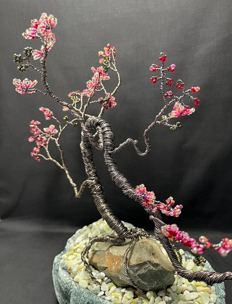 K&ouml;nnte beinhalten: Eine Drahtskulptur eines Bonsai-Baums mit rosa und gr&uuml;nen Perlen, die Bl&uuml;ten darstellen. Der Baum ist in einem grauen Steinsockel mit wei&szlig;en Kieselsteinen verwurzelt.