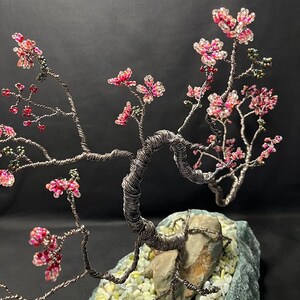 K&ouml;nnte beinhalten: Eine Drahtskulptur eines Bonsai-Baums mit rosa und wei&szlig;en Perlen, die Bl&uuml;ten darstellen. Der Baum steht auf einem grauen Steinsockel mit wei&szlig;en Steinen.