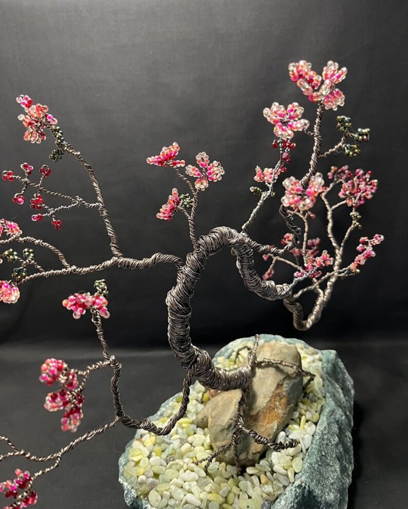 K&ouml;nnte beinhalten: Eine Drahtskulptur eines Bonsai-Baums mit rosa und wei&szlig;en Perlen, die Bl&uuml;ten darstellen. Der Baum steht auf einem grauen Steinsockel mit wei&szlig;en Steinen.