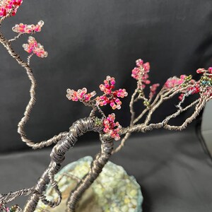K&ouml;nnte beinhalten: Eine Drahtskulptur eines Baumes mit rosa und wei&szlig;en Perlen, die Bl&uuml;ten darstellen. Der Baum steht in einem grauen und gr&uuml;nen Steintopf.