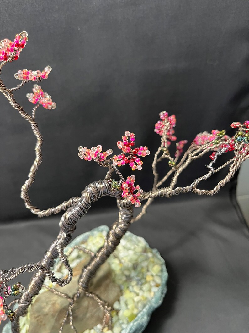 K&ouml;nnte beinhalten: Eine Drahtskulptur eines Baumes mit rosa und wei&szlig;en Perlen, die Bl&uuml;ten darstellen. Der Baum steht in einem grauen und gr&uuml;nen Steintopf.