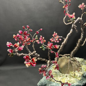 K&ouml;nnte beinhalten: Ein Bonsai-Baum aus Draht mit rosa und roten Perlen, die Bl&uuml;ten darstellen. Der Baum steht auf einem grauen Stein mit wei&szlig;en Steinen drumherum.