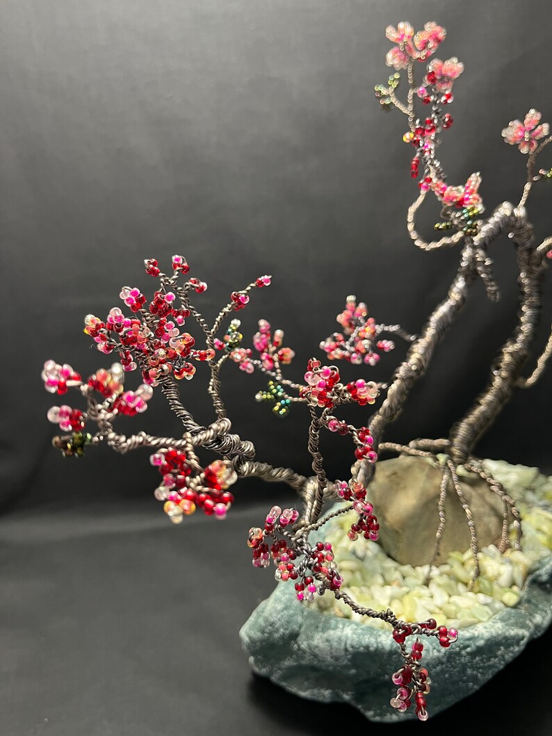 K&ouml;nnte beinhalten: Ein Bonsai-Baum aus Draht mit rosa und roten Perlen, die Bl&uuml;ten darstellen. Der Baum steht auf einem grauen Stein mit wei&szlig;en Steinen drumherum.