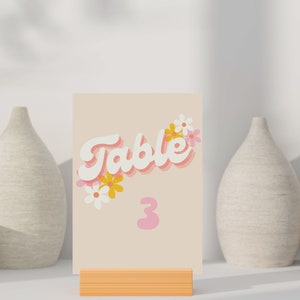 May include: A pink and white table number card with the text "Table 3" in a retro font. The card is decorated with white and yellow flowers.