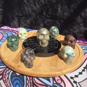 May include: Seven carved stone skull figurines, each with a different colour and pattern, arranged in a circle on a wooden tray. The skulls are made of green, brown, grey, and white stone. The tray has a black circular design in the centre.